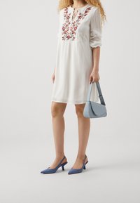 Vit klänning med blommönsterbroderi, långa ärmar och knytning vid halsen. Ljusblå handväska och blå slingback-klackar med spetsig tå.