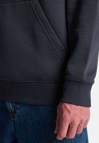 Close-up of a person wearing a dark sweatshirt with ribbed cuff and blue denim jeans, showing the lower torso and right hand.