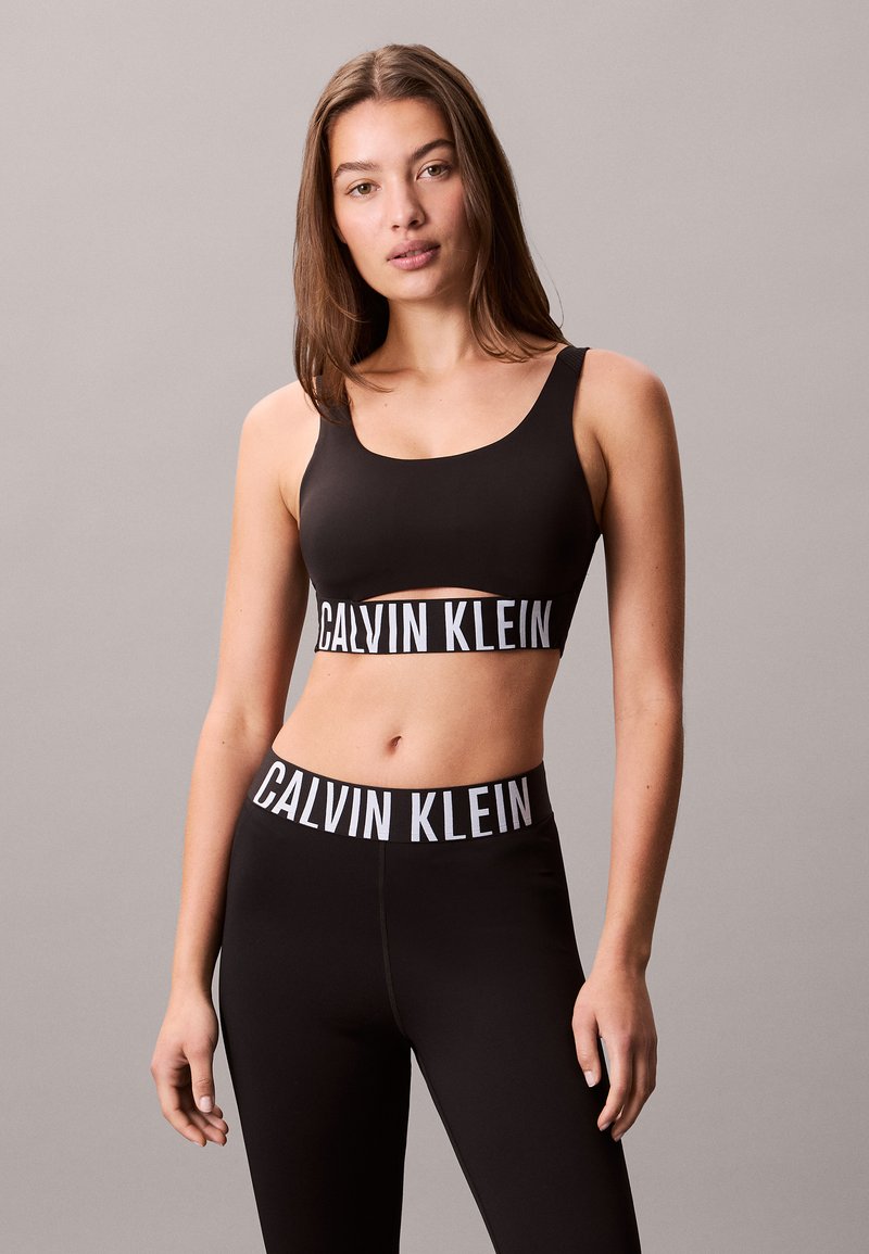 Black sports bra with a wide white "CALVIN KLEIN" logo band. Paired with matching black leggings featuring an identical logo waistband.