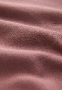 Teksturowana tkanina w miękkim różowym odcieniu, z subtelnymi falami i zdobieniami włókien. Materiał wydaje się gładki z jednolitą powierzchnią.