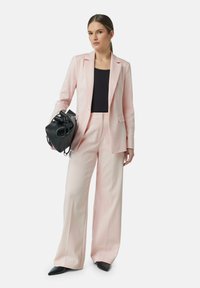 Completo su misura rosa chiaro con pantaloni a gamba larga, una giacca aderente e una maglia nera. Accessorio include una borsa nera con chiusura a coulisse.