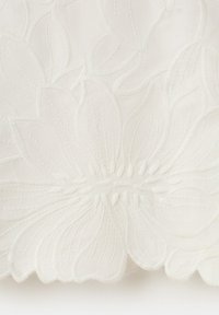 Tissu blanc avec broderie florale en relief présentant de grandes pétales et des détails texturés dans un motif répétitif.