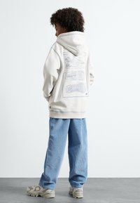 Krēmveida hoody ar zilu apavu grafiku aizmugurē, apvienots ar gaiši ziliem korduroja biksēm un pelēkiem sporta apaviem ar teksturētu zoli.