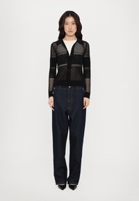 Black knitted cardigan with a dotted pattern, button closure, and long sleeves, paired with loose dark denim trousers and pointed black shoes.