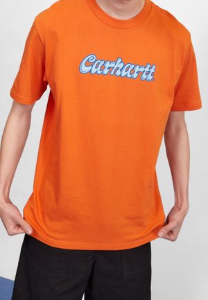 Persona que lleva una camiseta de manga corta de color naranja brillante con el logo "Carhartt" en azul y blanco en el pecho, sujetando el dobladillo de la camiseta.