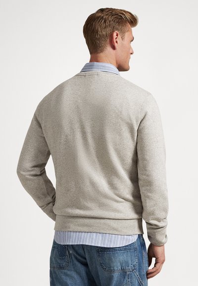 Ανοιχτό γκρι πλεκτό πουλόβερ με στρογγυλή λαιμόκοψη, ριμπ μανσέτες και τελείωμα. Φορεμένο πάνω από ένα μπλε και λευκό ριγέ πουκάμισο με γιακά, εμφανιζόμενο από πίσω.