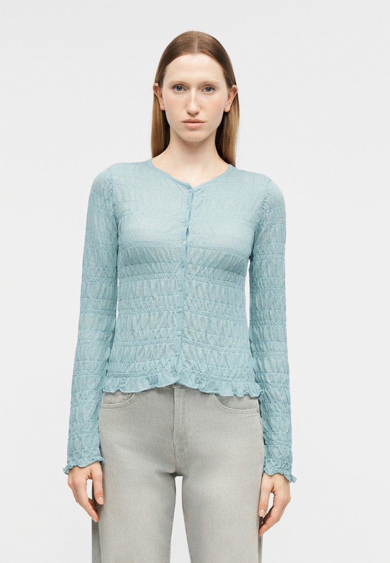 Γυναίκα που φοράει ανοιχτό μπλε υφασμάτινο ζακέτα με κουμπιά, μακριά μανίκια και βολάν στις άκρες, σε συνδυασμό με ανοιχτό γκρι ψηλόμεσο παντελόνι.