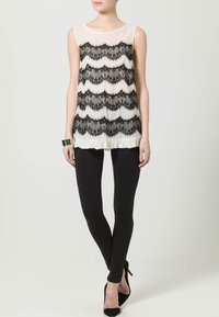 Zwart-wit mouwloos topje met een geschulpte kantdesign boven een doorschijnende stof, gecombineerd met zwarte leggings en hoge hakken.