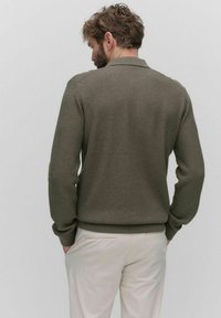 Olivgrön texturerad tröja med ribbad krage och manschetter, som har en snug passform och ett subtilt mönster, tillsammans med ljusa byxor.