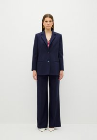 Sada s tmavě modrým blazerem a širokými kalhotami, s propracovaným střihem, rovnými kapsami a jednokolečkovým zapínáním. Doplněno špičatými podpatky.