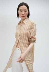 Beige knäppt skjorta med vita vertikala ränder, uppvikta ärmar och en ficka. Matchas med vita byxor. Slät tyg, avslappnad passform.