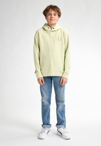 Felpa con cappuccio verde chiaro, dotata di tasca frontale e piccolo logo, abbinata a jeans in denim blu e sneakers bianche. Tessuto morbido.