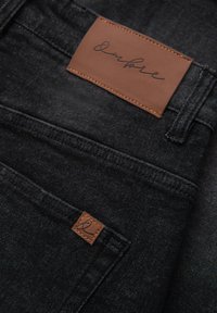 Primer plano de unos vaqueros de mezclilla negros que muestran dos parches de cuero marrón de la marca con logotipos en cursiva cerca de la cintura y el bolsillo.