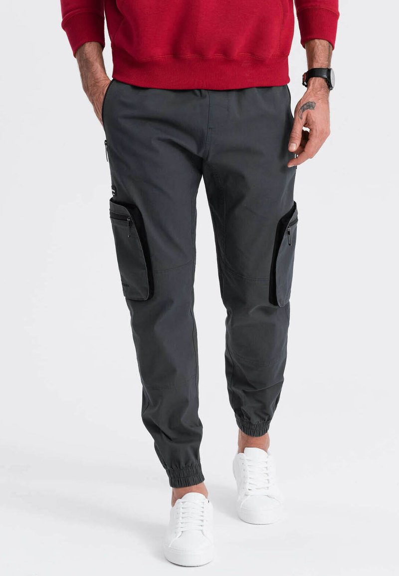 Cargo-Hosen in Dunkelgrau, mit Reißverschlusstaschen, elastischen Bündchen und einer glatten Textur. Kombiniert mit weißen Sneakern und einem roten Sweatshirt.