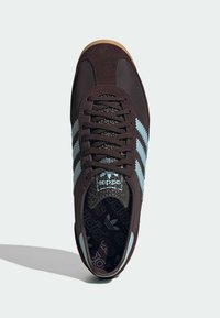 Καφέ σουέντ αθλητικό παπούτσι με ανοιχτό μπλε ρίγες, μαύρη εσωτερική σόλα με υφή και καφέ λαστιχένια σόλα. Διαθέτει στρογγυλά κορδόνια και κλασικό λογότυπο.