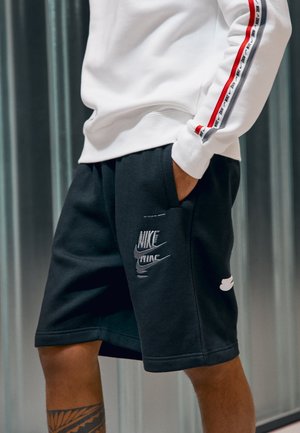 Musta shortsit sivutaskuilla, joissa on Nike-logopainatus ja valkoinen kontrastivärinen logo. Yhdistetty valkoisen collegepuseron kanssa.