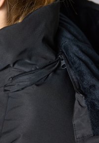 Jachetă neagră cu un guler înalt, având un buzunar cu fermoar și o căptușeală moale, texturată. Externul este elegant și neted.