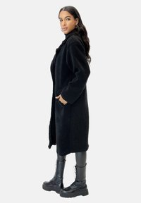 Schwarzer, langer, flauschiger Mantel mit Taschen, offenem Vorderteil und weiten Ärmeln, kombiniert mit schwarzen Schnürstiefeln, die einen vielschichtigen, strukturierten Look erzeugen.