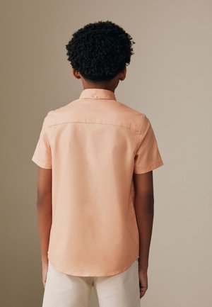 Chlapec s krátkymi kučeravými vlasmi, oblečený v svetlo broskyňovej košeli s krátkym rukávom a svetlých nohaviciach, otočený chrbtom k neutrálnemu pozadiu.