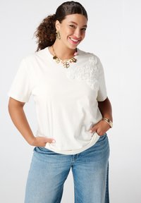 T-shirt blanc à manches courtes avec des embellissements floraux texturés d'un côté, associé à un jean large bleu clair et des bijoux dorés.
