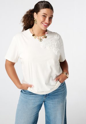 Witte t-shirt met korte mouwen en textuur van bloemrijke versieringen aan één kant, gecombineerd met lichtblauwe wijde jeans en gouden sieraden.