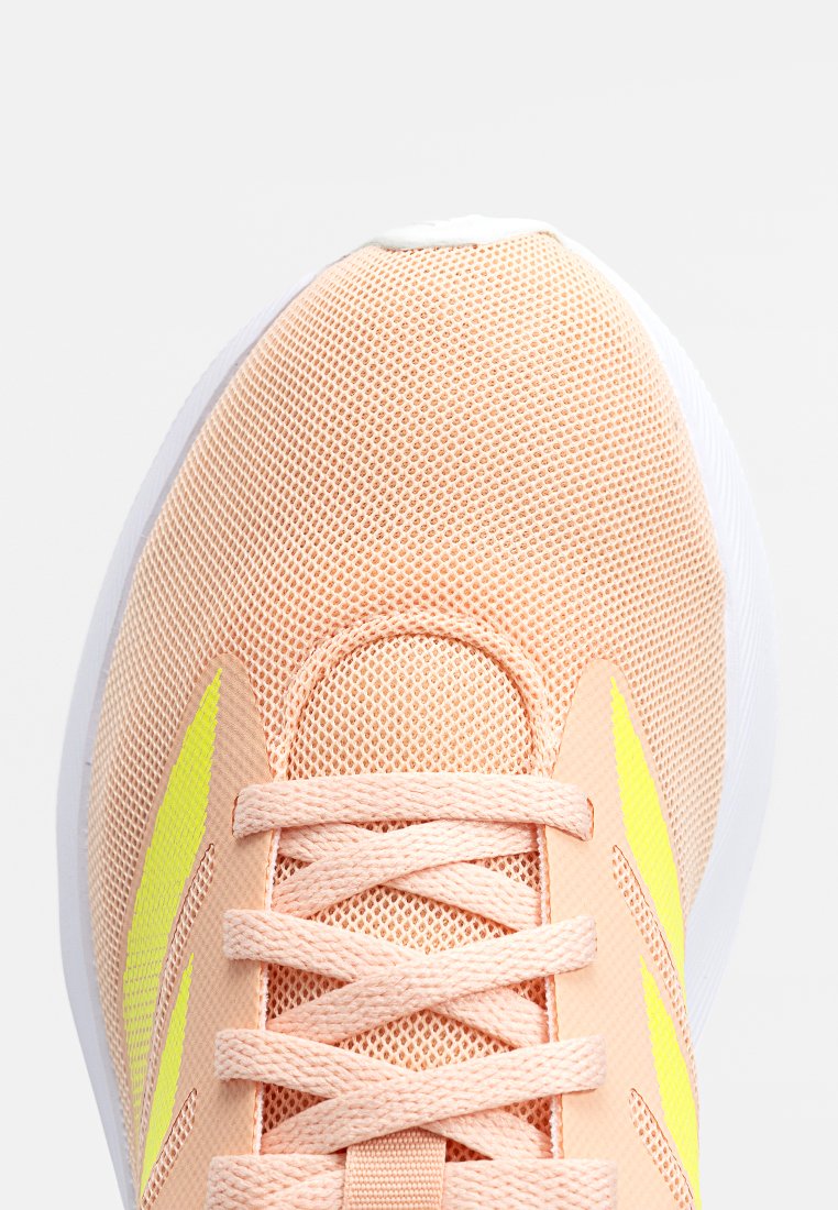 Zapatilla deportiva con una parte superior de malla en melocotón claro, acentos amarillos, cordones planos y una suela blanca acolchada. Presenta un diseño de punta redondeada.