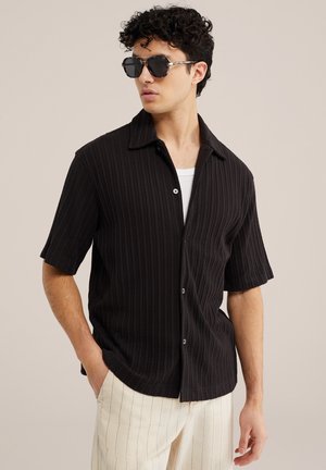 Jeune homme portant une chemise noire à manches courtes côtelée avec boutons, un tee-shirt blanc en dessous, un pantalon blanc à fines rayures et des lunettes de soleil noires angulaires, debout contre un mur beige.