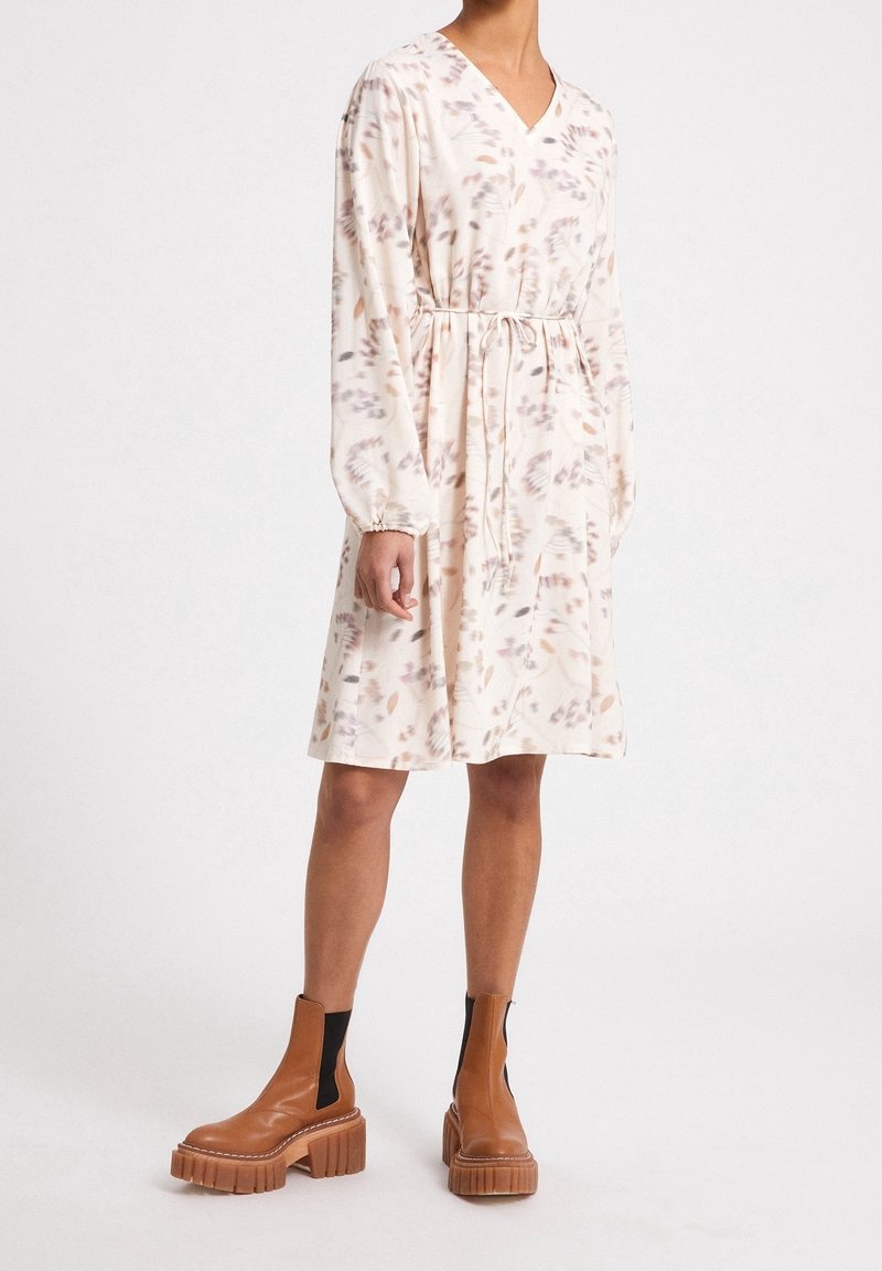 Vestido fluido de color crema con escote en V, mangas largas y cintura ajustada. Presenta un patrón floral en tonos apagados y puños fruncidos.