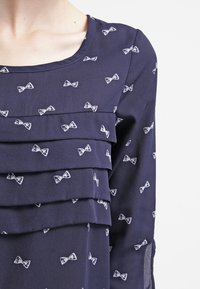 Blouse marine avec des volants superposés, ornée d'un petit motif de nœuds blancs. Fabriquée en tissu léger avec une texture lisse et un col rond.