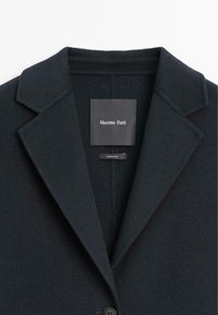Cappotto in misto lana di un elegante blu scuro, con rever a punta, chiusura con un singolo bottone e etichetta del marchio all'interno del colletto. Tessuto morbido.