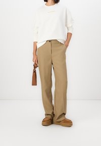Pantalones de pierna ancha beige con cierre de botón, combinados con una sudadera color crema que tiene un logo naranja. Bolsa marrón y zapatos beige gruesos.