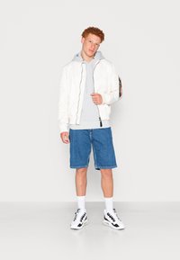 Giacca bomber bianca, felpa grigia chiara, pantaloni corti in denim, sneaker bianche con dettagli neri, in piedi davanti a uno sfondo neutro.