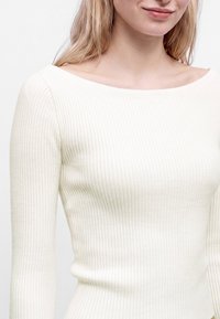 Jeune femme portant un haut moulant blanc côtelé à manches longues avec un large col rond sur un fond uni.