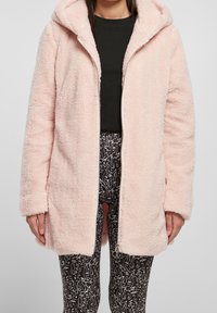 Abrigo con capucha rosa y esponjoso, con cierre frontal y bolsillos laterales. Presenta una textura suave y un diseño holgado. Lleva encima de una blusa negra y leggings con estampado.
