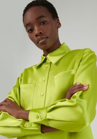 Camisa de satén verde lima con cuello abotonado, dos bolsillos frontales y mangas con puños. Textura suave y corte a medida.
