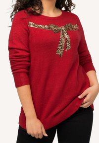 Czerwony sweter dziany z długimi rękawami, z detalem w postaci złotej cekinowej kokardy na piersi oraz ściągaczami na mankietach i u dołu. Tkanina o fakturze.