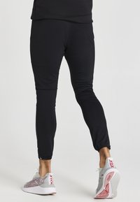 Černé sportovní kalhoty se tapered střihem, vyrobené z elastického materiálu, s rozepínacími manžetami a hladkým povrchem. V kombinaci se světlými teniskami.