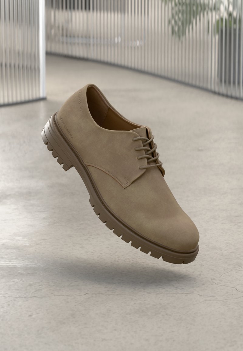 Beige lace-up men's shoe with thick treaded sole, hovering above textured concrete floor with vertical metal bars in the background.