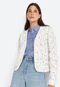 Weißer strukturierter Blazer mit mehrfarbigen Sprenkeln, mit rundem Halsausschnitt und offenem Vorderteil. Kombiniert mit einem blau-weiß gestreiften Hemd.