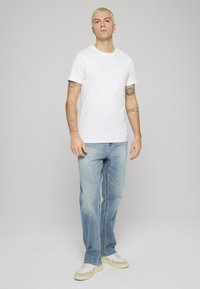 Camiseta blanca, jeans rectos azul claro y zapatillas beige. La modelo está de pie frente a un fondo gris neutro, mostrando un conjunto casual.