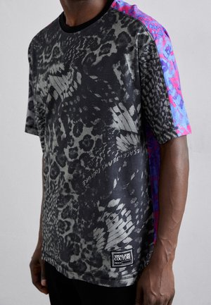 Mann trägt ein Versace Jeans Couture T-Shirt mit schwarzem und grauem Tiermuster auf der Vorderseite und kurzärmeligem lila-pink gemustertem Stoff vor einfarbigem Hintergrund.