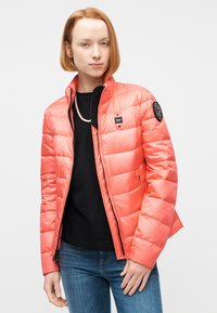 Femme aux cheveux courts roux portant une veste matelassée corail, une chemise noire, un collier de perles et un jean bleu, debout devant un fond blanc.