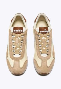 Scarpe sportive beige e crema con materiale scamosciato e tessuto, caratterizzate da un design unico dei lacci a incrocio e accenti marroni sul tallone e sul logo.