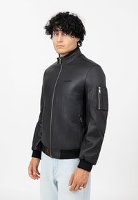 Giacca bomber nera in pelle con colletto alto, chiusura con zip e tasche laterali. Presenta polsini a coste e una tasca sulla manica con motivo a griglia.