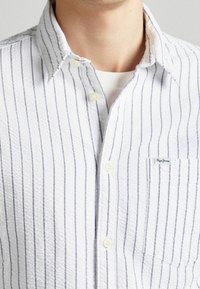 Camisa blanca con rayas verticales en azul marino, tela texturizada, cierre frontal con botones, cuello y un bolsillo en el pecho con una etiqueta de marca.