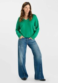 Groene v-hals trui met een losse pasvorm, gecombineerd met brede blauwe jeans. De outfit heeft een gestructureerd breisel en een casual denimstijl.