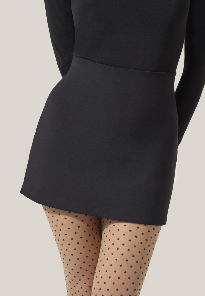 Kvinna med en svart minikjol och genomskinliga prickiga strumpbyxor, tillsammans med en figurnära svart långärmad topp mot en neutral bakgrund.