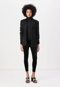 Blazer nero sartoriale con spalle strutturate, indossato sopra un dolcevita nero, abbinato a leggings neri e sandali con tacco massiccio.