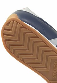 Semelle de chaussure avec motif de bande de roulement en zigzag beige et logo Adidas visible, fixée à une tige en cuir bleu marine et blanc.