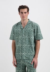 Korte mouwen shirt van groene stof met een getextureerd, bloemmotief. Inclusief een camp-col en knoopsluiting. Bijpassende broek zichtbaar.